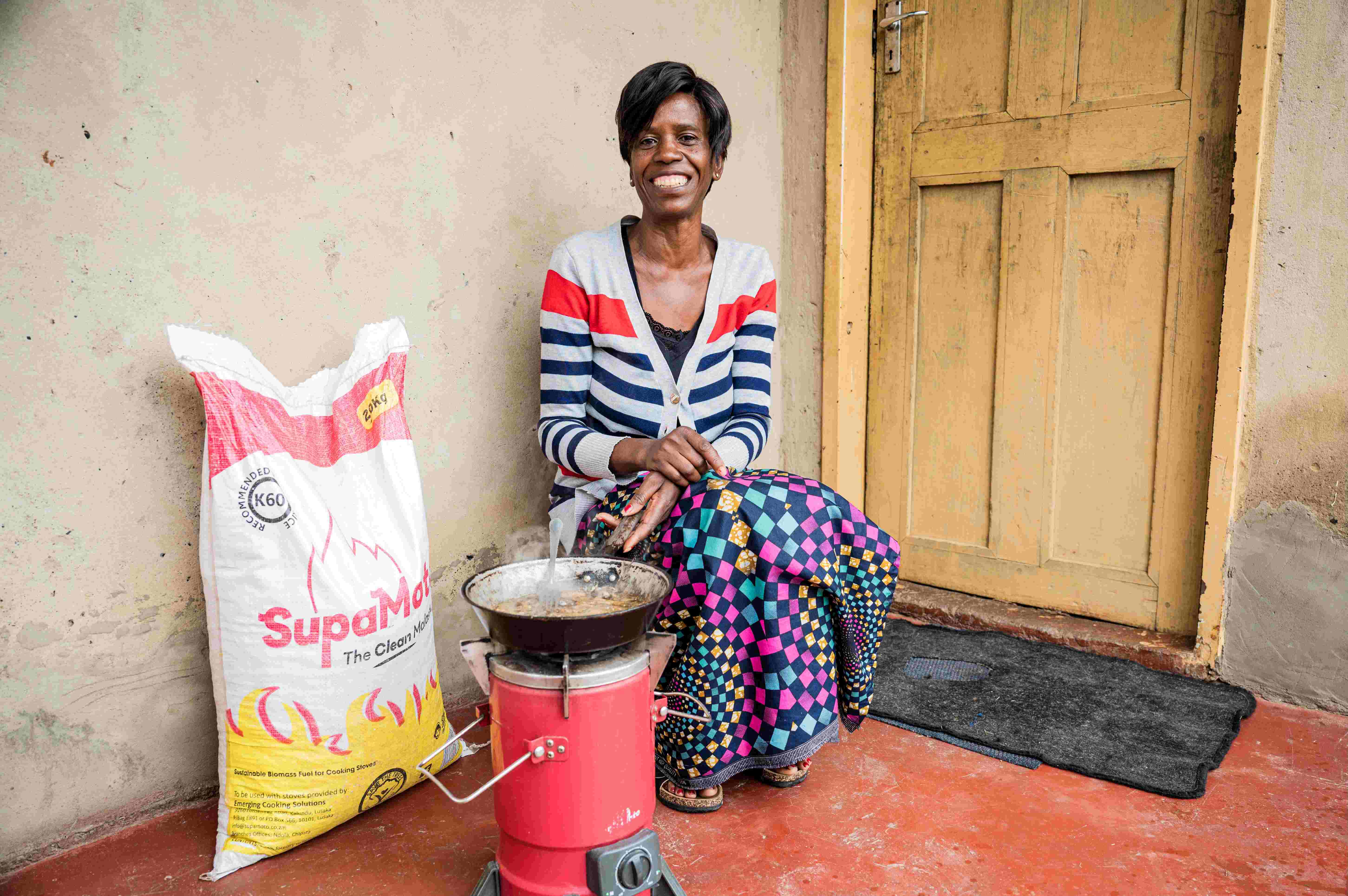 Woman sitting and smiling behind her clean cookstove and pellets by Emerging Cooking Solutions.