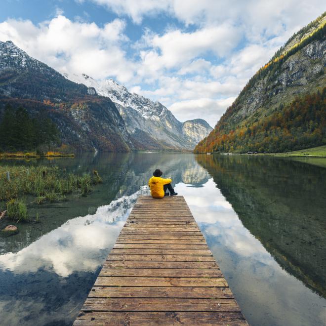 A man, wearing a yellow jacket and hat, is sitting alone on a boat pier admiring the Konigssee lake, Bavaria, Germany.