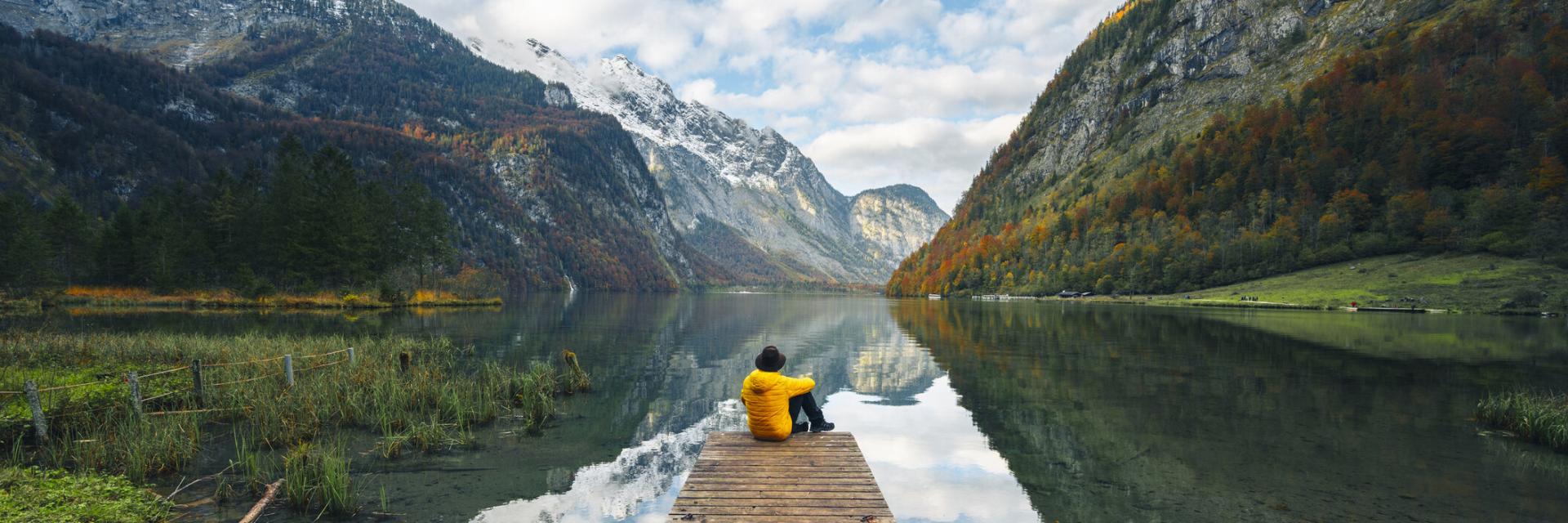 A man, wearing a yellow jacket and hat, is sitting alone on a boat pier admiring the Konigssee lake, Bavaria, Germany.